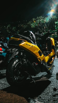 A yellow motorcycle is parked on a dimly lit street. The vehicle has a sleek design with the brand name 'NMax' visible on the side. It is positioned near some greenery, and there are other vehicles nearby. The scene is captured at night with artificial lighting creating a reflective effect on the bike's surface.