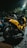A yellow motorcycle is parked on a dimly lit street. The vehicle has a sleek design with the brand name 'NMax' visible on the side. It is positioned near some greenery, and there are other vehicles nearby. The scene is captured at night with artificial lighting creating a reflective effect on the bike's surface.