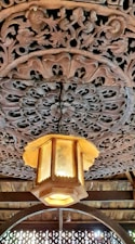 A detailed wooden carving decorates the ceiling with intricate floral patterns. Hanging from the center is an ornate lantern with a warm glow, made of wood and frosted glass, adding charm to the setting.