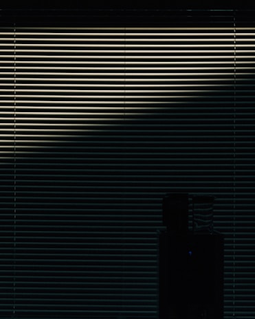 Horizontal blinds are partially open, allowing a dim light to pass through, casting a pattern of lines across the scene. In the lower part of the image, a silhouetted object is visible, possibly a piece of furniture or appliance.
