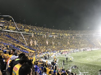 A lively scene at a packed football stadium, with a large crowd of enthusiastic supporters wearing yellow and blue. The stands are filled with banners and flags, and confetti is scattered on the field. The atmosphere is festive and celebratory as fans cheer and wave flags, and security personnel are visible along the perimeter of the field.