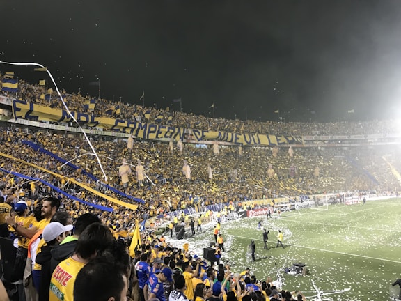 A lively scene at a packed football stadium, with a large crowd of enthusiastic supporters wearing yellow and blue. The stands are filled with banners and flags, and confetti is scattered on the field. The atmosphere is festive and celebratory as fans cheer and wave flags, and security personnel are visible along the perimeter of the field.