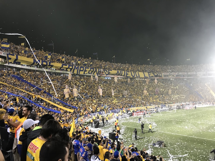 A lively scene at a packed football stadium, with a large crowd of enthusiastic supporters wearing yellow and blue. The stands are filled with banners and flags, and confetti is scattered on the field. The atmosphere is festive and celebratory as fans cheer and wave flags, and security personnel are visible along the perimeter of the field.