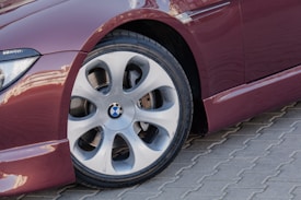 A close-up of a red car's wheel featuring a BMW logo in the center, set on a brick-patterned pavement. The car has a glossy finish and the tire appears clean and new.