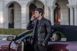 A person wearing a dark leather jacket stands next to a convertible car with the door open, talking on a phone. The background features a building with large arches and columns, suggesting an urban or sophisticated setting.