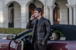 A person wearing a dark leather jacket stands next to a convertible car with the door open, talking on a phone. The background features a building with large arches and columns, suggesting an urban or sophisticated setting.