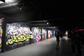 A dimly lit underground passageway featuring vibrant graffiti art on the walls. Several people are casually walking or standing, observing the art. The environment has a slightly industrial feel with exposed beams and overhead lighting.