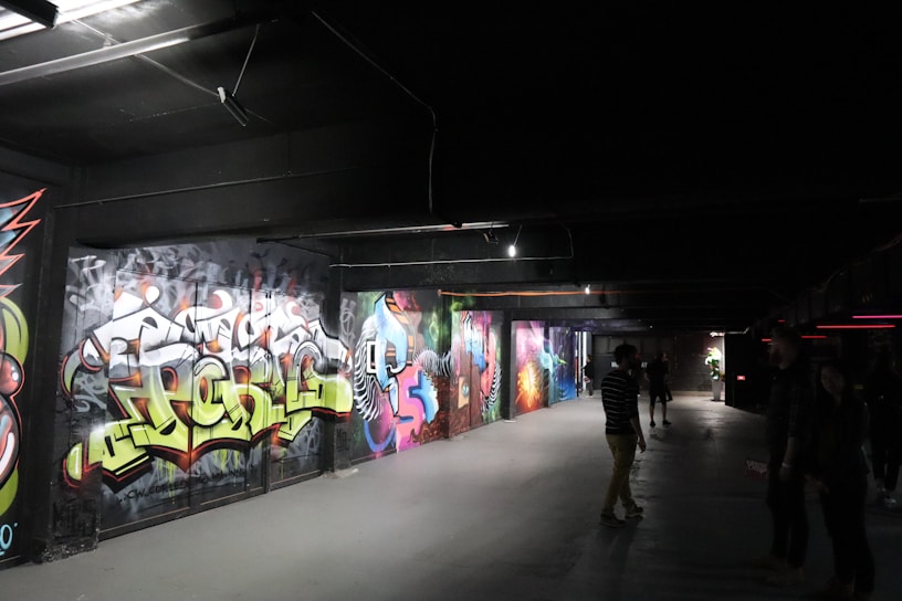 A dimly lit underground passageway featuring vibrant graffiti art on the walls. Several people are casually walking or standing, observing the art. The environment has a slightly industrial feel with exposed beams and overhead lighting.