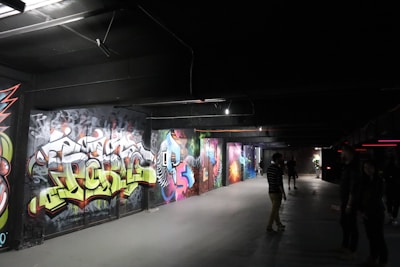 A dimly lit underground passageway featuring vibrant graffiti art on the walls. Several people are casually walking or standing, observing the art. The environment has a slightly industrial feel with exposed beams and overhead lighting.