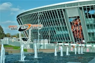 A modern stadium with a large glass facade and an electronic billboard displaying an athlete in action. In front, there are fountains spouting water against a backdrop of a clear blue sky. The name 'Donbass Arena' is prominently displayed on a metal structure.