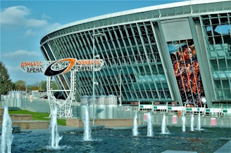 A modern stadium with a large glass facade and an electronic billboard displaying an athlete in action. In front, there are fountains spouting water against a backdrop of a clear blue sky. The name 'Donbass Arena' is prominently displayed on a metal structure.