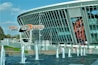 A modern stadium with a large glass facade and an electronic billboard displaying an athlete in action. In front, there are fountains spouting water against a backdrop of a clear blue sky. The name 'Donbass Arena' is prominently displayed on a metal structure.