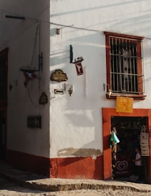A sunlit corner of a rustic building with white and red walls. There are vintage signs and a barred window above a small shop entrance. The shop door is open, revealing a person standing inside and a child nearby. Newspapers are for sale at the entrance.