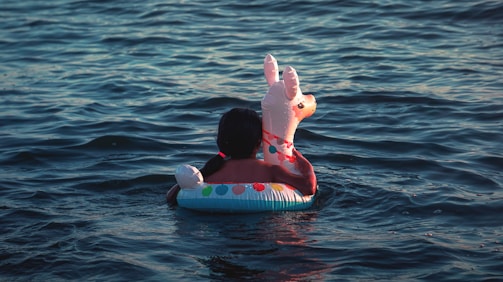 A person is sitting in a colorful inflatable pool toy shaped like an animal, floating in a body of water. The water appears calm with gentle ripples, and the lighting suggests it is either early morning or late afternoon.