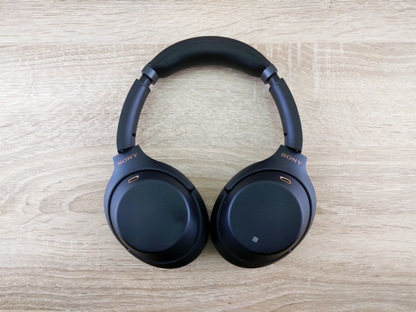 A pair of black over-ear headphones is placed on a light wood surface. The headphones have a sleek design with padded ear cups and a comfortable headband. The brand logo is visible on the side.