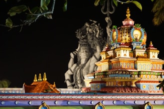 A vibrant, intricately decorated temple tower stands in front of a large, detailed statue. The temple structure is adorned with colorful patterns and embellishments, featuring shades of blue, red, yellow, and green. In the background, a large gray statue holds prominent religious significance, adding a sense of grandeur and spirituality to the scene. The dark night sky emphasizes the vivid colors and details of the temple architecture.