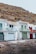 A row of three small seaside houses with brightly colored doors and balconies stands by the water's edge. Each house is painted white with blue, green, and red accents. The rocky hillside behind adds a natural backdrop. A person in summer clothing is walking along the waterfront carrying a bag, giving the scene a relaxed and leisurely atmosphere.