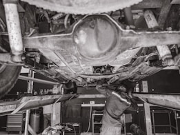 A mechanic is working underneath a vehicle that's elevated on a hydraulic lift. The scene captures various auto parts and tools scattered around the workshop. The environment appears to be a busy auto repair shop.