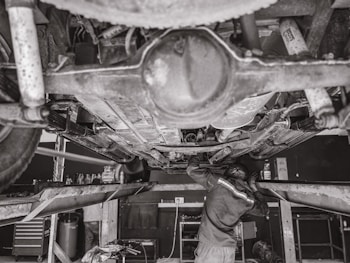 A mechanic is working underneath a vehicle that's elevated on a hydraulic lift. The scene captures various auto parts and tools scattered around the workshop. The environment appears to be a busy auto repair shop.