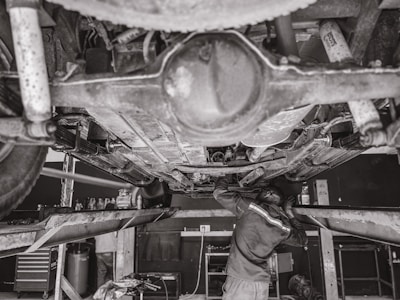 A mechanic is working underneath a vehicle that's elevated on a hydraulic lift. The scene captures various auto parts and tools scattered around the workshop. The environment appears to be a busy auto repair shop.