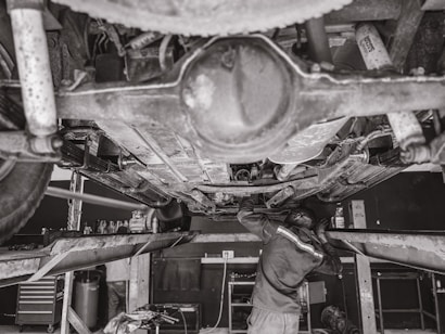 A mechanic is working underneath a vehicle that's elevated on a hydraulic lift. The scene captures various auto parts and tools scattered around the workshop. The environment appears to be a busy auto repair shop.