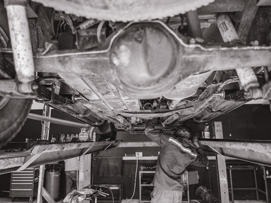 A mechanic is working underneath a vehicle that's elevated on a hydraulic lift. The scene captures various auto parts and tools scattered around the workshop. The environment appears to be a busy auto repair shop.