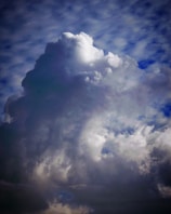 A large, towering cloud formation with varying shades of white and gray against a partly cloudy sky. The cloud is dense and billowing, suggesting an impending storm or heavy rainfall. Light and shadows create a dramatic contrast, emphasizing the cloud's three-dimensional texture.