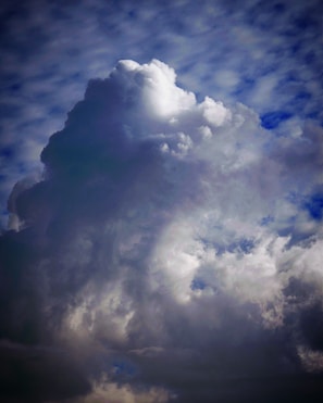 A large, towering cloud formation with varying shades of white and gray against a partly cloudy sky. The cloud is dense and billowing, suggesting an impending storm or heavy rainfall. Light and shadows create a dramatic contrast, emphasizing the cloud's three-dimensional texture.