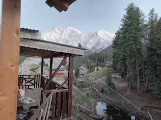 A wooden cabin porch overlooks a picturesque landscape featuring snow-capped mountains and dense forests. Small rustic houses are scattered across the scene, and a narrow stream runs through the grassy clearing surrounded by trees.