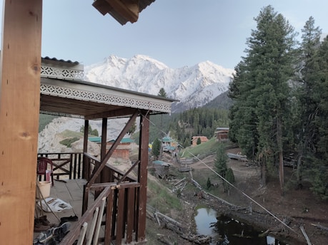 A wooden cabin porch overlooks a picturesque landscape featuring snow-capped mountains and dense forests. Small rustic houses are scattered across the scene, and a narrow stream runs through the grassy clearing surrounded by trees.