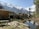 A scenic mountainous landscape featuring a wooden cabin on the left surrounded by a grassy area and scattered wooden planks. A small pond reflects the clear blue sky and adjacent tall pine trees. Snow-capped mountains dominate the background, creating a stunning natural setting.