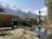 A scenic mountainous landscape featuring a wooden cabin on the left surrounded by a grassy area and scattered wooden planks. A small pond reflects the clear blue sky and adjacent tall pine trees. Snow-capped mountains dominate the background, creating a stunning natural setting.