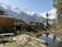 A scenic mountainous landscape featuring a wooden cabin on the left surrounded by a grassy area and scattered wooden planks. A small pond reflects the clear blue sky and adjacent tall pine trees. Snow-capped mountains dominate the background, creating a stunning natural setting.