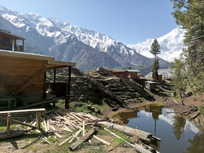 A scenic mountainous landscape featuring a wooden cabin on the left surrounded by a grassy area and scattered wooden planks. A small pond reflects the clear blue sky and adjacent tall pine trees. Snow-capped mountains dominate the background, creating a stunning natural setting.