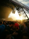 A large crowd of people gathered at an outdoor music festival during sunset, with the stage structure visible and lit by the sun's rays. Concertgoers are closely packed together, some wearing hats and sunglasses.