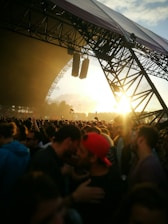 A large crowd of people gathered at an outdoor music festival during sunset, with the stage structure visible and lit by the sun's rays. Concertgoers are closely packed together, some wearing hats and sunglasses.