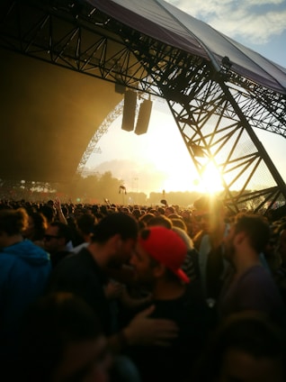 A large crowd of people gathered at an outdoor music festival during sunset, with the stage structure visible and lit by the sun's rays. Concertgoers are closely packed together, some wearing hats and sunglasses.