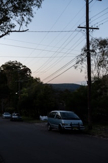 A quiet suburban street lined with trees and telephone poles. Overhead, numerous power lines crisscross the sky, which shows a gradient from light to darker blue as the evening sets in. Several cars are parked along the side of the street, leading towards a distant hill covered in bushland.
