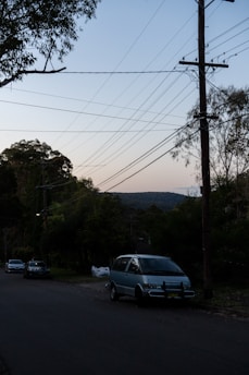 A quiet suburban street lined with trees and telephone poles. Overhead, numerous power lines crisscross the sky, which shows a gradient from light to darker blue as the evening sets in. Several cars are parked along the side of the street, leading towards a distant hill covered in bushland.