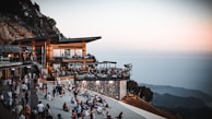 A modern, multi-level outdoor restaurant perched on a mountainside, bustling with people enjoying the scenic view. The backdrop consists of expansive mountain vistas under a subtly colored sky transitioning into evening. Visitors are gathered on a wide terrace, with some sitting on the ground, creating a lively and relaxed atmosphere.