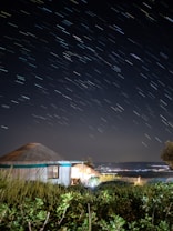 A long exposure night photograph capturing star trails in the sky above a yurt set amidst lush greenery. The sky is dark, with trails of light from the stars creating a dynamic effect. A distant view of city lights is visible on the horizon, adding a sense of depth and contrast to the natural surroundings.