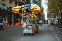 A hot dog stand is positioned on a city sidewalk with yellow and blue umbrellas bearing the brand name 'Sabrett.' It's surrounded by various items for sale, including water bottles and snacks. The street features buildings under construction, traffic cones, vehicles, and a few pedestrians walking by.