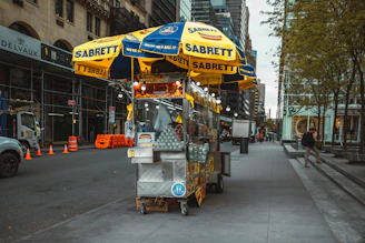 Rows of Sabrett hotdogs grilling over an open flame, with city buildings blurred in the background.