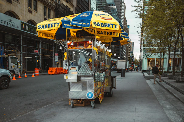 Rows of Sabrett hotdogs grilling over an open flame, with city buildings blurred in the background.