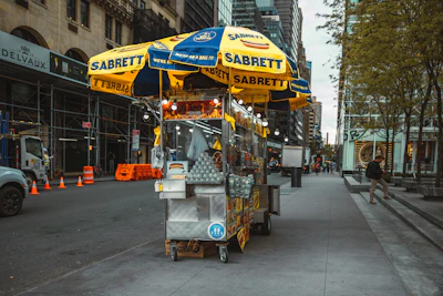 A hot dog stand is positioned on a city sidewalk with yellow and blue umbrellas bearing the brand name 'Sabrett.' It's surrounded by various items for sale, including water bottles and snacks. The street features buildings under construction, traffic cones, vehicles, and a few pedestrians walking by.