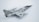 A model aircraft with a sleek, metallic design, featuring jet engines and distinctive military markings. The aircraft has dual wings and pointed nose with noticeable detail in its construction. The backdrop is a plain, light-colored surface that highlights the aircraft's shiny surface.