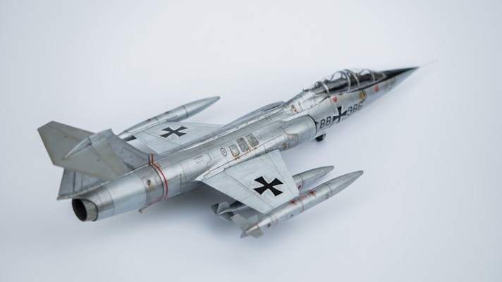 A model aircraft with a sleek, metallic design, featuring jet engines and distinctive military markings. The aircraft has dual wings and pointed nose with noticeable detail in its construction. The backdrop is a plain, light-colored surface that highlights the aircraft's shiny surface.