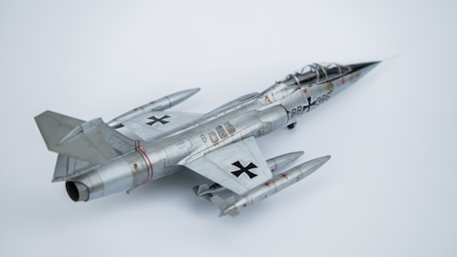 A model aircraft with a sleek, metallic design, featuring jet engines and distinctive military markings. The aircraft has dual wings and pointed nose with noticeable detail in its construction. The backdrop is a plain, light-colored surface that highlights the aircraft's shiny surface.