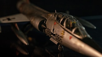 A detailed model of a military aircraft with a focus on its cockpit and nose section. The aircraft features a distinctive emblem and numbering, indicative of a specific air force. It is depicted in a dimly lit setting, highlighting the sleek metallic surface and intricate details of the model.
