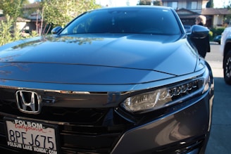 A sleek, shiny gray Honda car is parked in a residential driveway. The front view shows the vehicle's headlights and grille, with a visible Folsom Lake Honda license plate. In the background are trees and the facade of a house, along with a person walking nearby.
